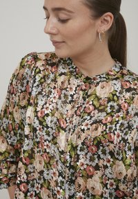 Blusa floreale con sfondo nero, caratterizzata da fiori rosa e bianchi. Realizzata in tessuto leggero e trasparente con scollo rotondo e dettaglio di bottoni.