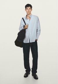 Camicia a righe blu e bianche con bottoni, jeans in denim scuro, scarpe nere e una borsa grigia con texture. Il modello si trova davanti a uno sfondo semplice.