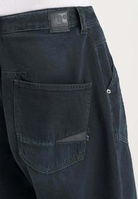 Zwarte denim broek met een achterzak met klep en een klein label, textuur op het oppervlak en een standaard tailleband.