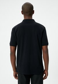 Svart polo-tröja med en texturerad krage, korta ärmar och rak nederkant. Tyget verkar slätt och lätt. Enkel design utan mönster.