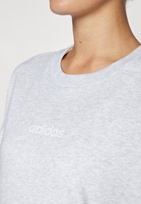 Svetlo šedá mikina so širokým výstrihom, jemnou textúrou a bielym logom "adidas" na prednej strane.