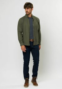 Vīrietis valkā olīvu zaļu pogātā krekla pārvalku virs tumši pelēka džempera, tumšās džinsās, brūnas jostas un brūnus ādas zābakus, stāv ar rokām atviegloti.