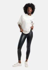 Witte oversized t-shirt met een kleine afbeelding, gecombineerd met zwarte glanzende leggings. Model draagt witte sneakers. Soepele textuur en strakke pasvorm.