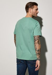 Camiseta de algodón verde menta con mangas cortas, escote de tripulación y un corte relajado, combinada con jeans azul oscuro. Detalle visible de tatuaje en el brazo.