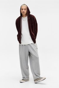 Mahagonový manžestrový hoodie s tkaničkami, bílým tričkem, světle šedými tepláky a vzorovanými teniskami. Model stojí s rukama v kapsách.