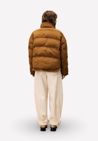 Persoon in een bruine gewatteerde jas, beige wijde broek met opgerolde pijpen en zwarte schoenen, staand met de rug naar de kijker op een effen achtergrond.