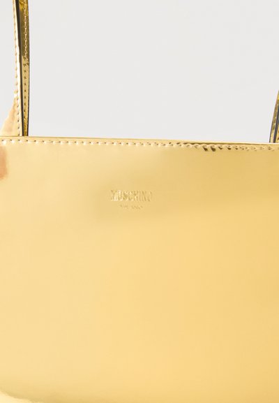 Λαμπερή χρυσή ορθογώνια τσάντα χειρός με λεπτές λαβές και ανάγλυφο λογότυπο «Moschino Milano» στο κέντρο μπροστά.