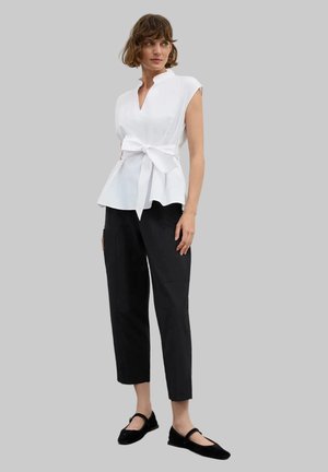 Donna che indossa una canotta bianca con fiocco frontale, pantaloni neri alla caviglia e scarpe Mary Jane nere, in piedi davanti a uno sfondo grigio.