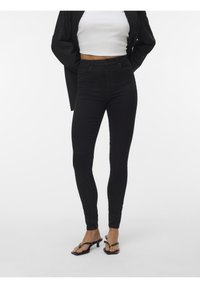 Femme portant un jean skinny taille haute noir, un top court blanc, un blazer noir et des sandales noires à talons, debout sur un fond blanc.