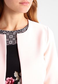 Różowa kurtka o minimalistycznym designie, z okrągłym dekoltem i gładką teksturą. Pod spodem czarna bluzka z akcentami w kwiatowy wzór.