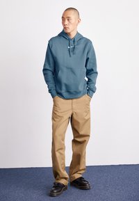 Sweatshirt com capuz em azul-teal, com um bolso frontal e logo branco, combinado com calças largas em bege e sapatos pretos com solas espessas.