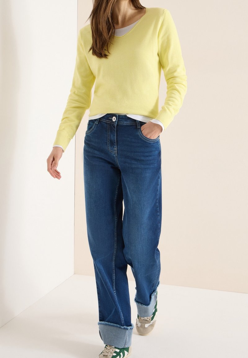 Femme portant un pull jaune, un sous-vêtement blanc, un jean bleu avec revers, et des baskets beige avec des bandes vertes, debout devant un fond crème.
