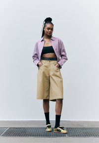 Lila strukturiertes Shirt, schwarzes Crop-Top, Leopardenmustergürtel, beige Shorts, goldene Sneakers und schwarze Socken. Stehend auf einer strukturierten Oberfläche.
