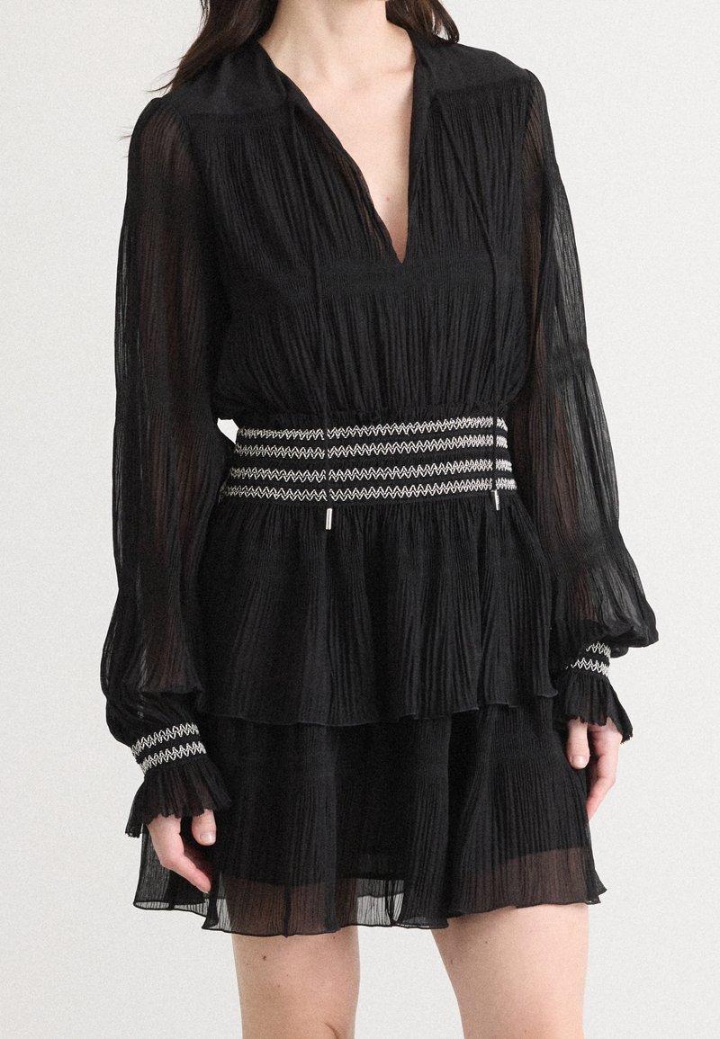 Robe noire plissée à manches longues, décolleté en V et taille cintrée. Caractérisée par un tissu texturé et une bande décorative rayée à la taille.