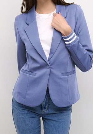 Femme portant un blazer bleu clair cintré avec des poignets rayés, une chemise blanche et un jean bleu, tenant le revers du blazer d'une main.