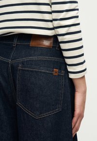Détail d'une personne portant un jean en denim bleu foncé avec des écussons en cuir de la marque et une chemise blanche à manches longues avec des rayures horizontales noires.