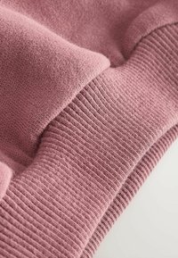 Ružová pletená látka s hladkou textúrou a žebrovanými akcentmi. Má mäkký, vrstvený vzhľad s rôznymi odtieňmi ružovej.