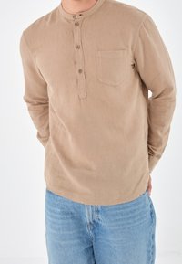Langsleeve, tan henley-shirt met een ronde halslijn, drie-knops sluiting en een enkele borstzak. Gedragen met lichtblauwe jeans.
