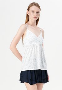 White floral top with thin straps, subtle lace trim, V-neck design, and gathered waist, paired with a layered navy skirt.