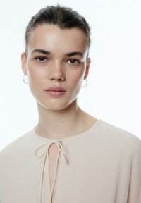 Beige Bluse mit rundem Halsausschnitt, Bindeschließe vorne, aus weichem Stoff. Das Model trägt dezentes Makeup und Ohrreifen.