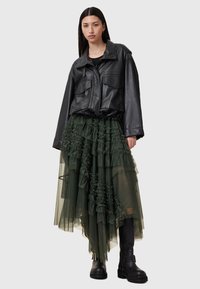 Chaqueta de cuero negra corta con bolsillos de parche, combinada con una falda de tul verde con capas y volantes. Botas negras hasta la rodilla completan el look.