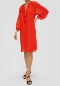 Rotes plissiertes Kleid mit einem Rüschendetail am Ausschnitt, langen Ärmeln und einer lockeren Passform, präsentiert mit schwarzen Riemchensandalen.