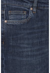 Jeans de denim azul oscuro con un bolsillo delantero, costuras sutiles y botones de cobre. La tela parece suave con una textura ligeramente utilizada.