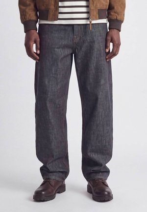 Homme portant un jean denim foncé et ample, des bottes en cuir marron à lacets, une veste marron et une chemise rayée noir et blanc, debout devant un fond uni.