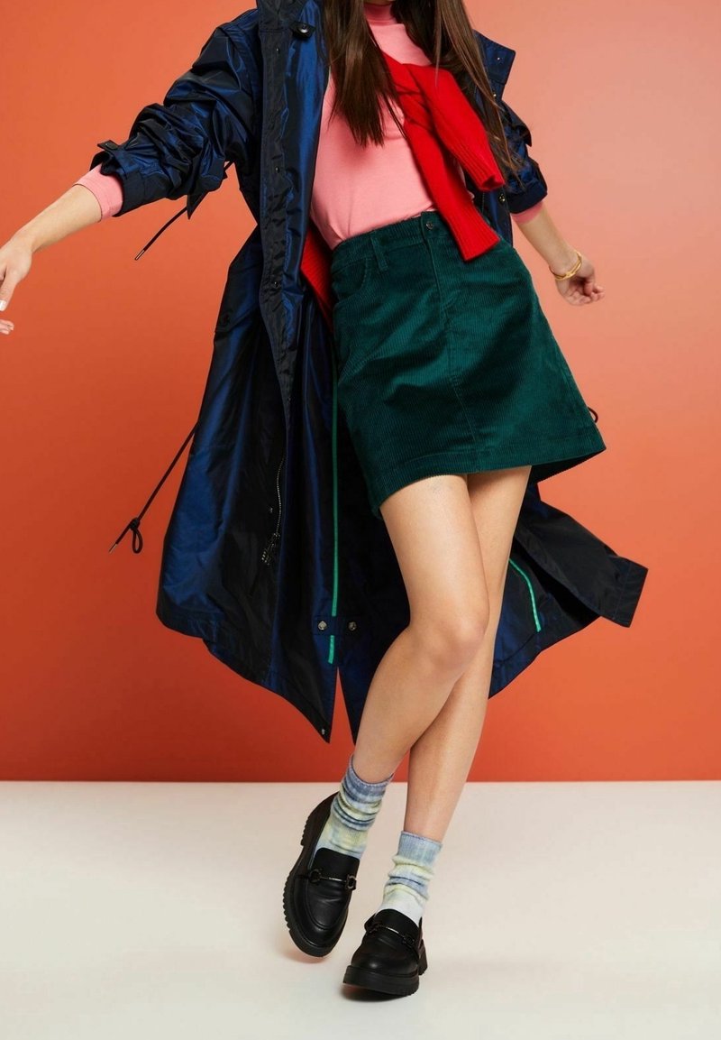 Marineblauwe regenjas, roze top en rode trui om de hals geknoopt; groene corduroy rok, gebloemde sokken en zwarte loafers maken de outfit compleet.