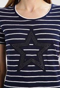 Camiseta de rayas azul marino con rayas blancas, con un gran diseño de estrella negra decorado con tachuelas de metal. Cuello redondo, mangas cortas.