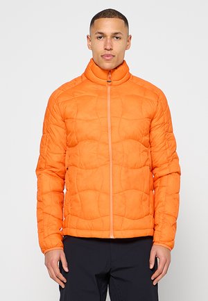 Mężczyzna w jasnopomarańczowej pikowanej kurtce z wysokim kołnierzem i czarnych spodniach, stojący na jednolitym jasnym tle.