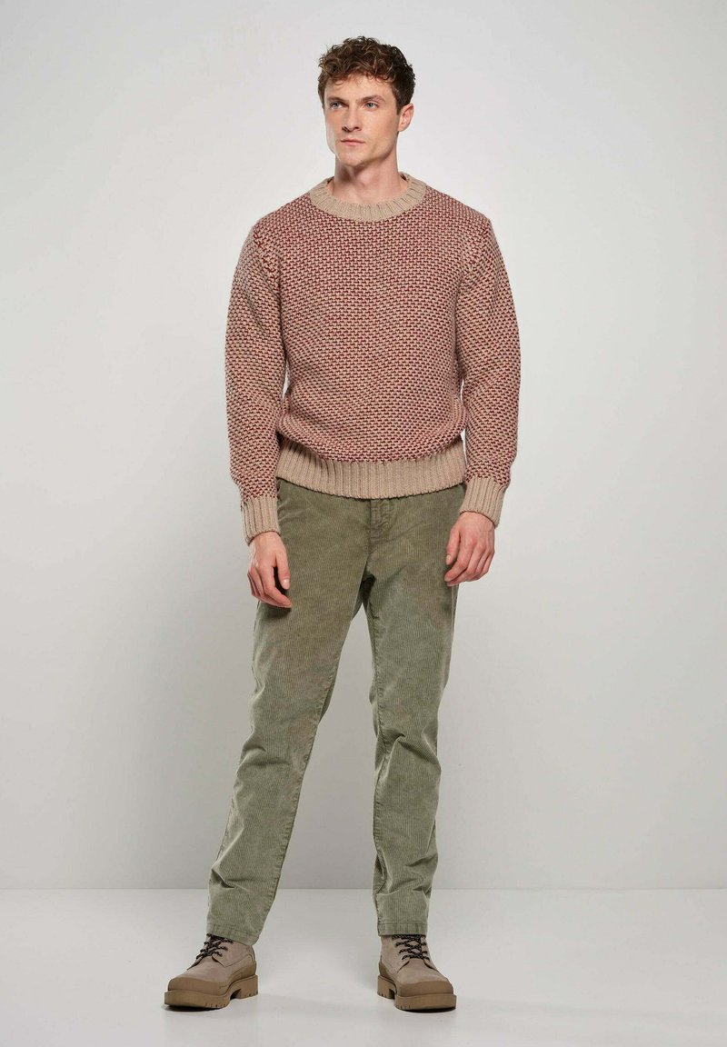Gebreide trui in rode en beige tinten, ronde hals; gecombineerd met lichtgroene corduroybroeken. Model draagt platte, lichte schoenen.