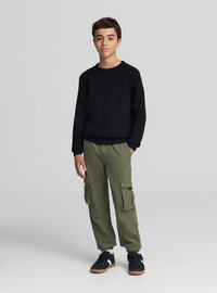 Maglione nero lavorato a maglia con trama, pantaloni cargo verde oliva con tasche laterali e sneakers nere con suola color cuoio. Outfit semplice e casual.