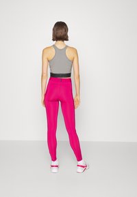 Una donna in piedi di spalle indossa un crop top grigio a spalle scoperte, leggings fuxia a vita alta, calzini bianchi e sneakers bianche con dettagli rosa.