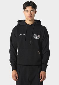 Černá mikina s kapucí vyrobená z bavlny s přední kapsou. Obsahuje bílé vyšívané logo a text. Ribované manžety a lem pro lepší přiléhavost.