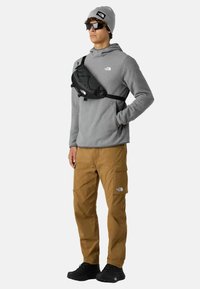 Szara polarowa bluza z kapturem, czarna torba na ramię, brązowe spodnie cargo, czarne sneakersy, szary beanie, okulary przeciwsłoneczne; posiada logo na torbie i odzieży.