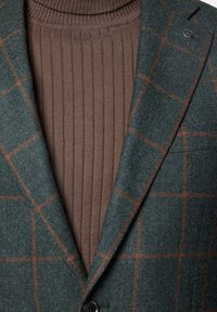 Zielona marynarka w kratę z teksturowanej tkaniny, nałożona na brązowy sweter z wysokim kołnierzem, z pojedynczym guzikiem i dopasowanym kołnierzykiem.