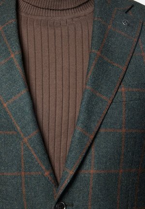 Zielona marynarka w kratę z teksturowanej tkaniny, nałożona na brązowy sweter z wysokim kołnierzem, z pojedynczym guzikiem i dopasowanym kołnierzykiem.