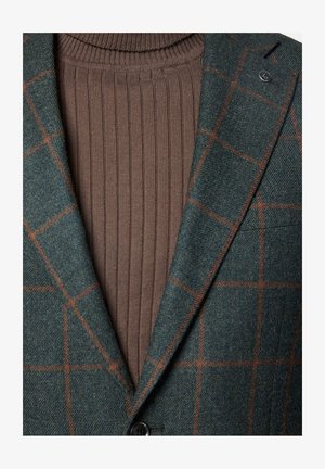 Zielona marynarka w kratę z teksturowanej tkaniny, nałożona na brązowy sweter z wysokim kołnierzem, z pojedynczym guzikiem i dopasowanym kołnierzykiem.