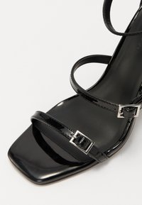 Sandale à talon haut en cuir verni noir avec trois brides incurvées et bout carré. Dispose de boucles en métal argenté sur les brides de cheville réglables.