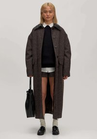 Lange bruine jas met een textuurpatroon, zwarte leren kraag, gecombineerd met een marineblauwe trui, shorts en zwarte loafers. Inclusief een zwarte tote bag.