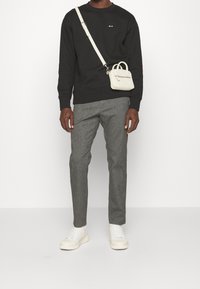 Muž v černé košili s dlouhým rukávem, šedých kalhotách, bílých teniskách a malé bílé crossbody tašce na jednobarevném bílém pozadí.