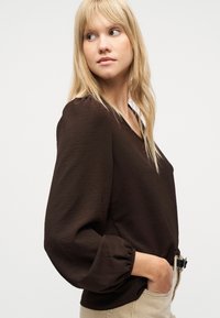 Ξανθιά γυναίκα με μακριά μαλλιά που φοράει μια φαρδιά σκούρα καφέ μπλούζα με μπαλόν μανίκια και μπεζ παντελόνι, κοιτάζοντας προς τα αριστερά της.