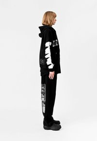 Sudadera negra con gráfico blanco en la manga, pantalones negros con acentos impresos y zapatillas negras chunky. Tela suave, corte relajado.