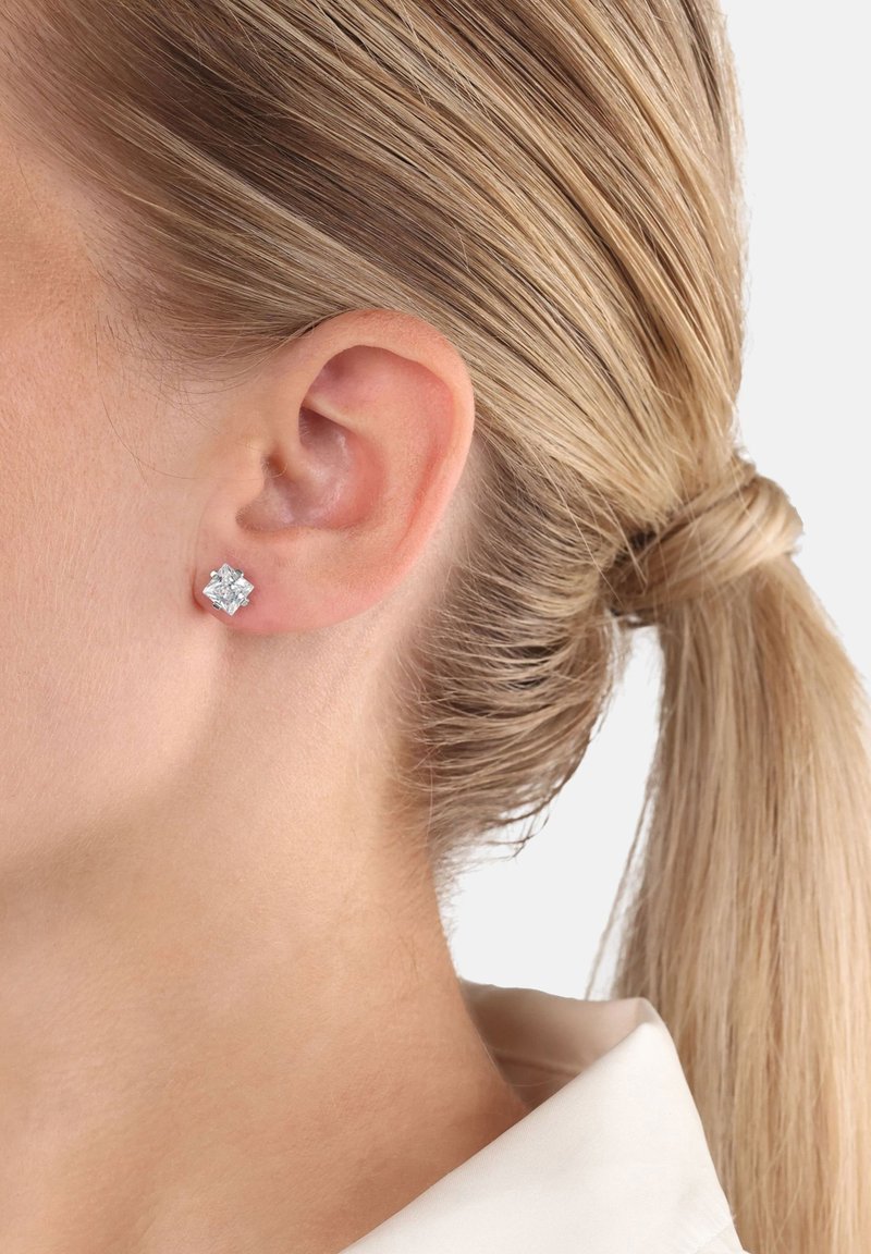 Mujer con cabello rubio recogido en una coleta, que lleva un pendiente de diamante en forma cuadrada en su oreja izquierda, sobre un fondo blanco.