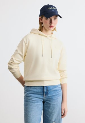 Fiatal nő krémszínű kapucnis pulóverben, világoskék farmerben, sötétkék sapkával, amelyen szöveg található, és ezüst függő fülbevalóval, egy kézzel a háta mögött áll.