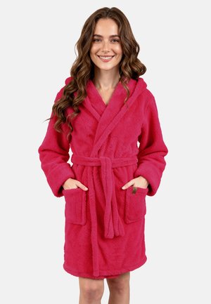 Donna con capelli lunghi e ricci che indossa un accappatoio in pile con cappuccio rosa acceso, con cintura e tasche frontali, sorridente con le mani nelle tasche.