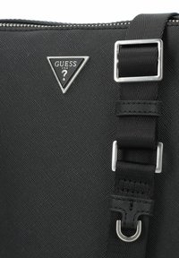 Bolsa mensajera negra con un acabado texturizado, herrajes plateados, correa ajustable y un logotipo triangular de Guess en el frente.
