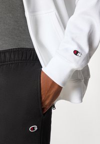 Grijze T-shirt met korte mouwen onder een witte sweater met rits en zwarte sweatpants. De sweatpants hebben een geborduurd logo nabij de zak.