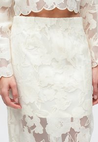 Witte kanten top met bloemenmotief en golvende randen, gecombineerd met een bijpassende transparante rok. De stof heeft ingewikkelde bloempatronen en texturen.